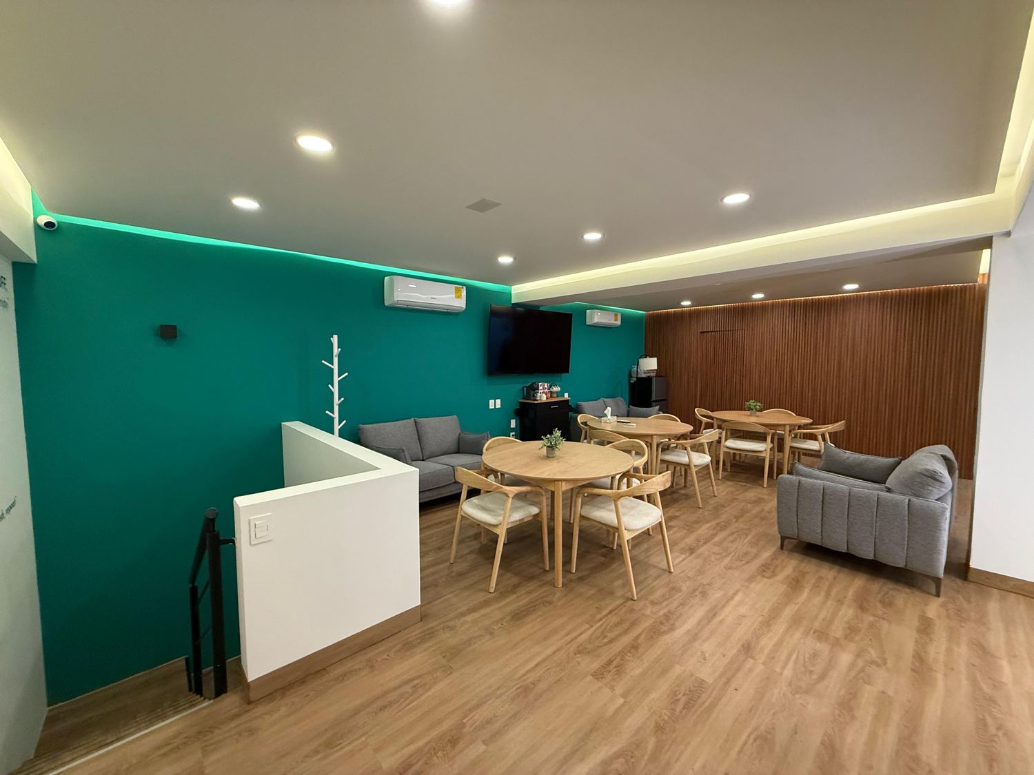 Sala de descanso de una clínica con muro verde, mesas redondas de madera, sillones grises y luz indirecta en el plafón
